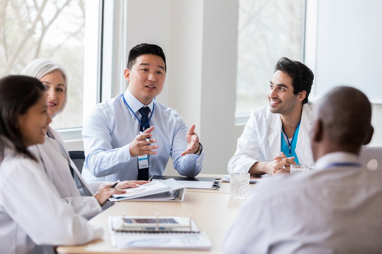 Young male pharmaceutical sales representative gestures while discussing a new medicine with doctors and other people in hospital management.